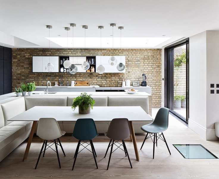 An open plan white kitchen with banquette seating, exposed brick walls and glass pendant lighting.