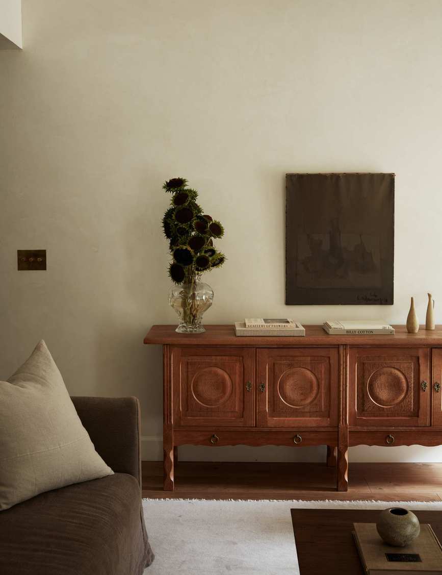 Mid-century wooden sideboard featuring a clear vase of sunflowers, a muted wall tapestry, and various collected objects