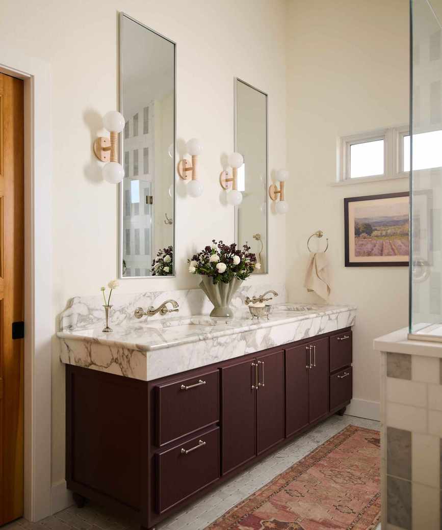 Bathroom with burgundy cabinetry, marble sink, marble floor, pink persian rug and two large mirrors with round sconces