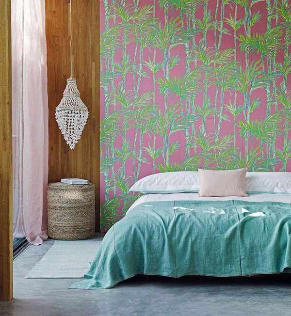 A colorful bedroom with a pink and green patterned statement wall, wood panelling, a macrame hanging, rattan stool, and a low bed with blue bedding.