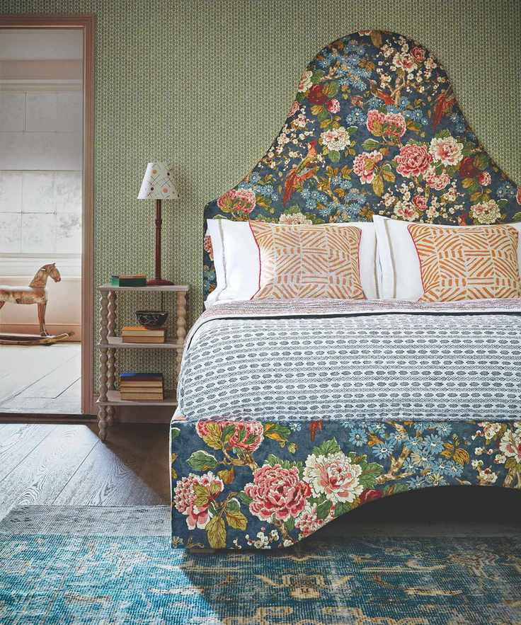 A green maximalist bedroom with a large floral patterned bed with statement headboard, patterned bedding, and orange patterned cushions. Beside the bed is a wooden bedside table with books and a wooden lamp with a white lampshade on top.
