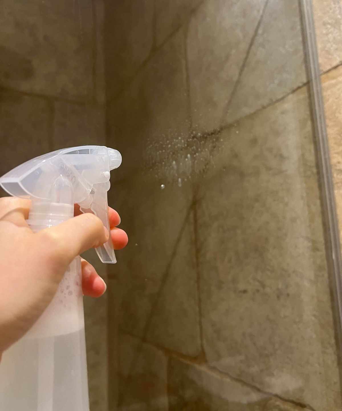 A hand spraying a shower screen with a clear spray bottle with neutral beige tiles behind
