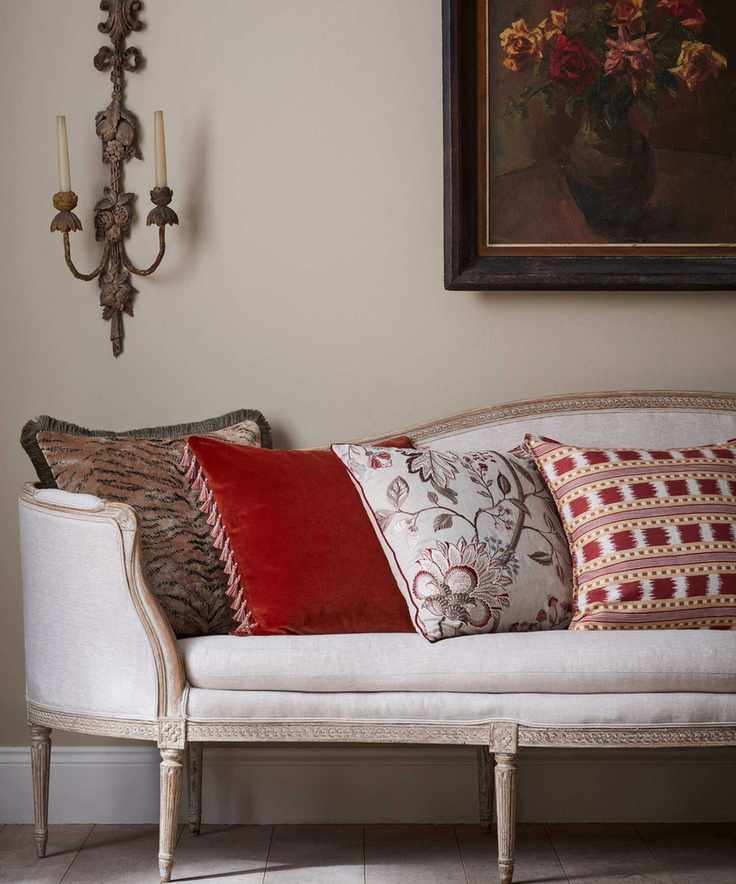 Vintage looking white upholstered chaise with four throw pillows in different patterns and designs in white, red, brown and taupe. There is an ornate wall-mounted candle holder with slim candles, and framed art on the wall with autumnal blooms