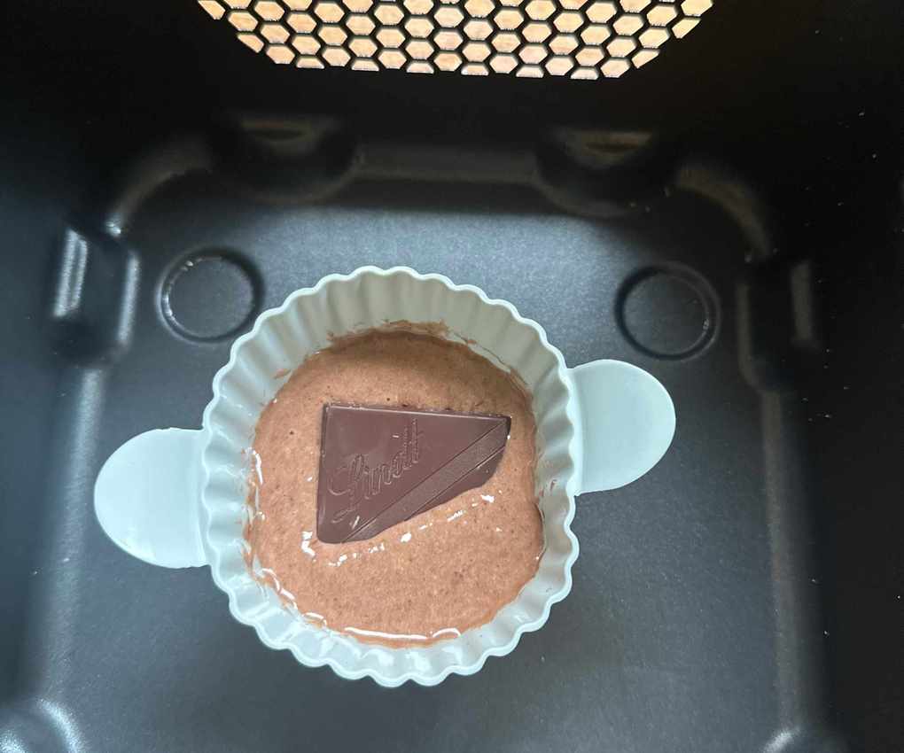 A small round blue silicone baking ramekin with chocolate cake batter and a square of chocolate inside, in a black air fryer basket
