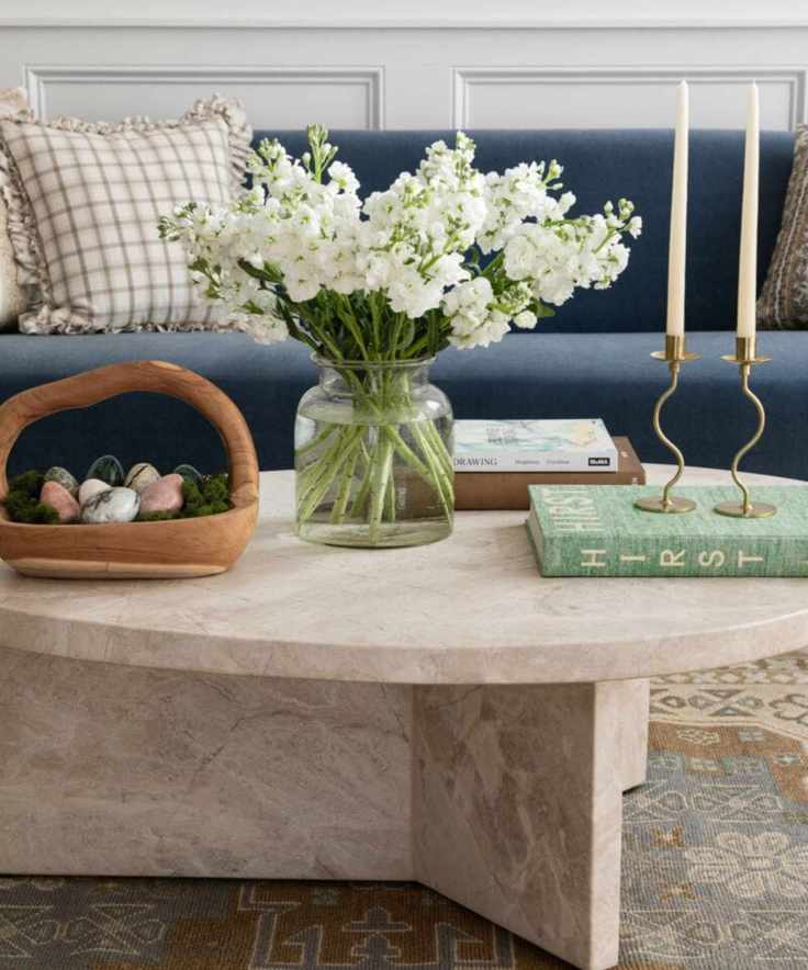 Living room with a navy blue couch and a marble round coffee table decorated for Easter with flowers and a wooden basket filled with decorative Easter eggs