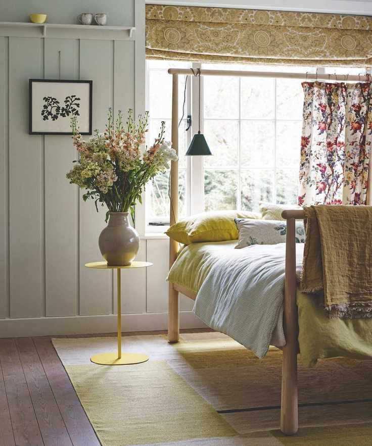 A bright bedroom with large windows, four poster bed, yellow bedding, yellow bedside table with large vase of flowers, yellow rug, and dark wood flooring. The walls are pale green panelled.