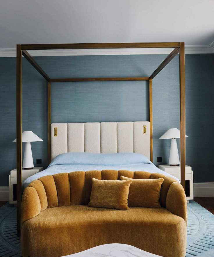 a blue grasscloth wallpapered primary bedroom with a four posted bed and mustard velvet sofa at the foot of the bed