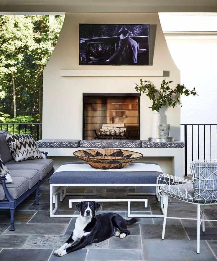 outdoor living area surrounded around a large outdoor working fireplace and wall mounted TV, with a tiled patio area with sofa, ottoman and chairs plus a black and white dog