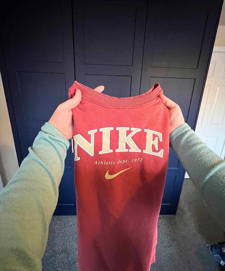 The image shows a point of view shot of a peachy-orange Nike long sleeve sweater being held in front of the camera with the arms now folded back in the air