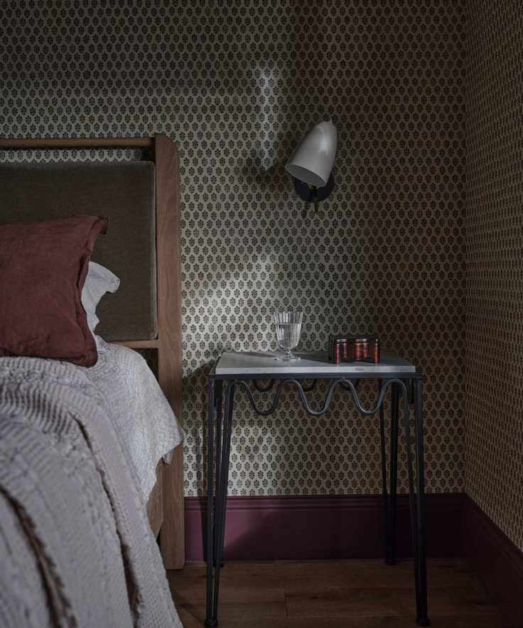 a small cottage bedroom with ditsy wallpaper and purple skirting boards, a wooden bed with green velvet headboard and a wavy metal nightstand