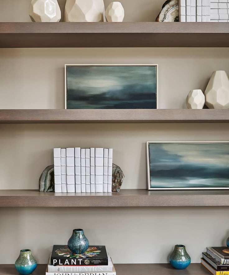 Wooden open shelving against a neutral painted wall. Blue abstract artwork in frames on the shelves beside white books between, help up with bookends