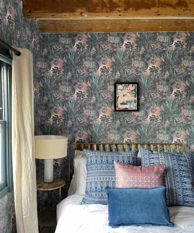 farmhouse style bedroom with blue and pink wallpaper, beams, blue and pink bed pillows, table lamp, cream drapes