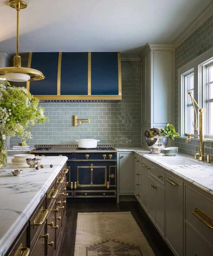 A large kitchen with light green cabinets, an island with a marble countertop, a dark blue oven and hood, pale green backsplash tiles, and a neutral runner rug on the dark flooring.