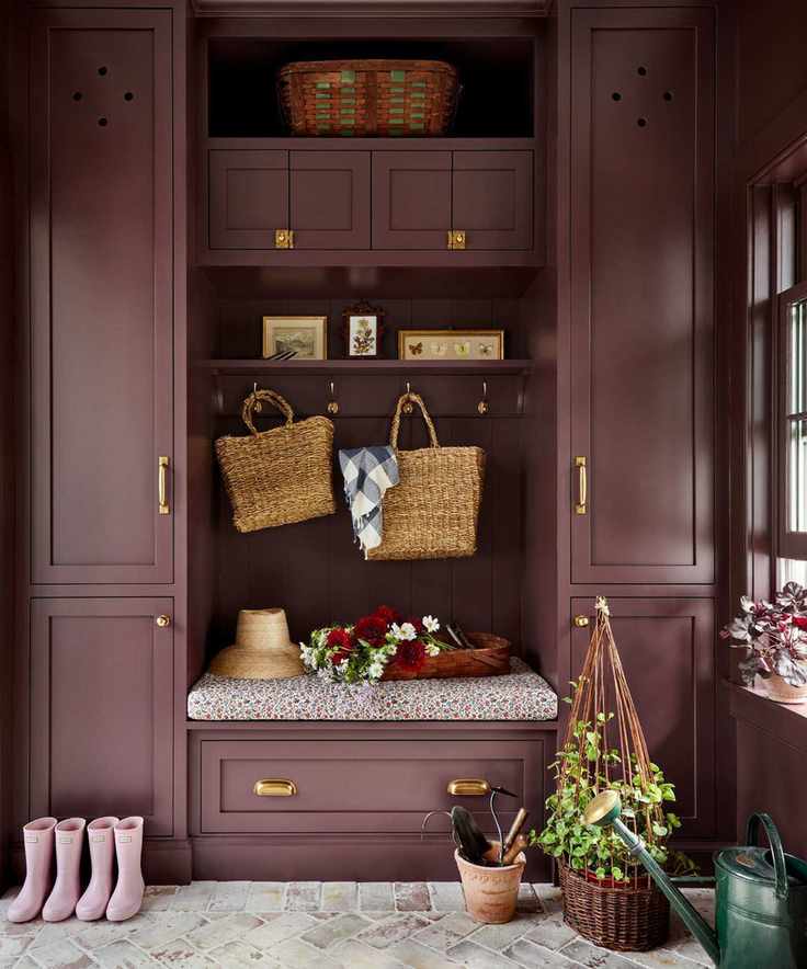 A mudroom with dark purple floor-to-ceiling cabinets with brass hardware, gray brick flooring, and coat hooks in the middle of the cabinets with bags, hats, and decorative objects.