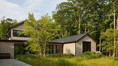 A modern shingle-style house with black-trimmed windows and a metal roof, surrounded by lush green grass and a dense forest.