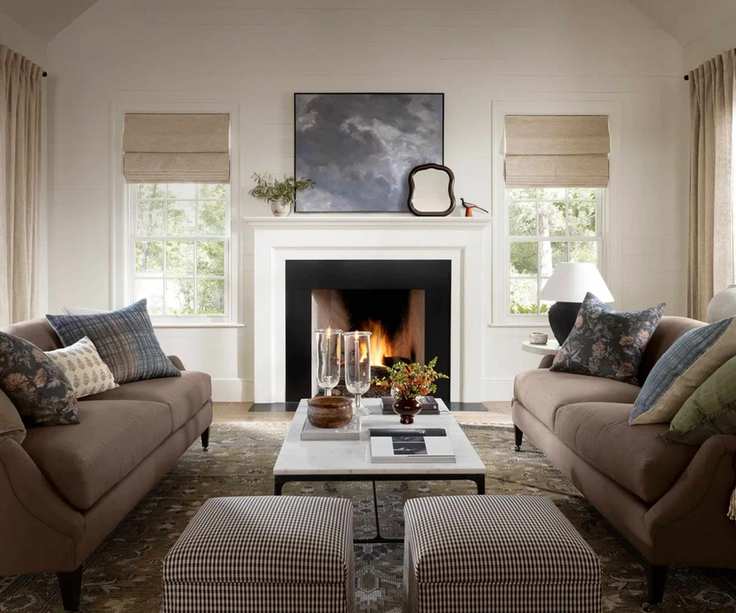 cozy neutral living room styled for fall using the mcgee and co fall 2025 collection. A large white fire is styled with a blue abstract painting, while two taupe sofas face each other styled with plaid and floral pillows and a marble coffee table with small plaid ottomans