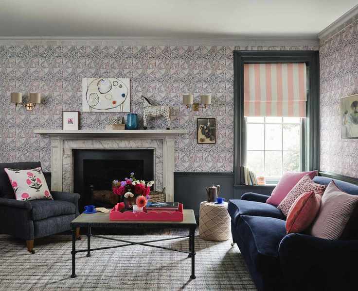 Living room with dado atelier wallpaper, blue painted paneling and wooden features, blue sofa and armchair, pink and red cushions, black metal coffee table, marble fireplace, pink and white striped blind, two shade wall lamps and wall art