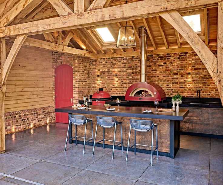 A covered outdoor kitchen with a red pizza oven, black countertops, and a wooden canopy