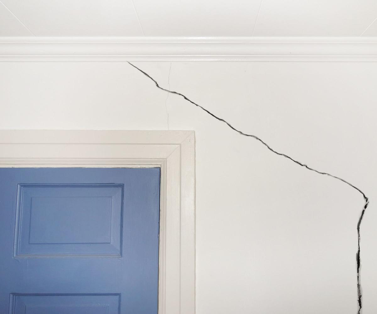 A large deep crack in a white wall above a white doorframe and blue door.