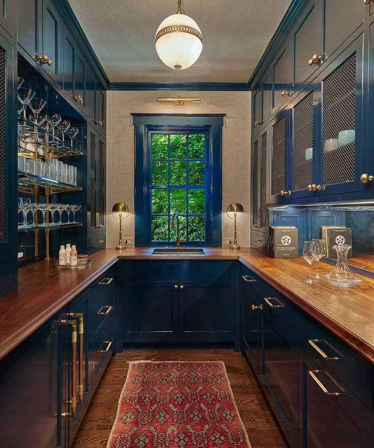 A kitchen with dark blue cabinets, wooden countertops, gold hardware, and a dark wood floor topped with a red Persian runner rug