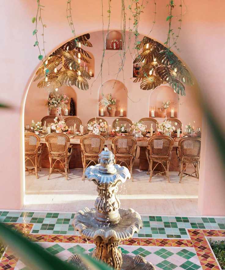 Courtyard dining set up with colorful floor tiles, pink walls and a stone fountain in the foreground and a table within an arch decorated with flowers in the background