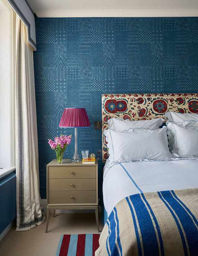 blue bedroom with blue wallpaper, patterned fabric headboard, blue stripe throw and bedding, bedside, pink lamp