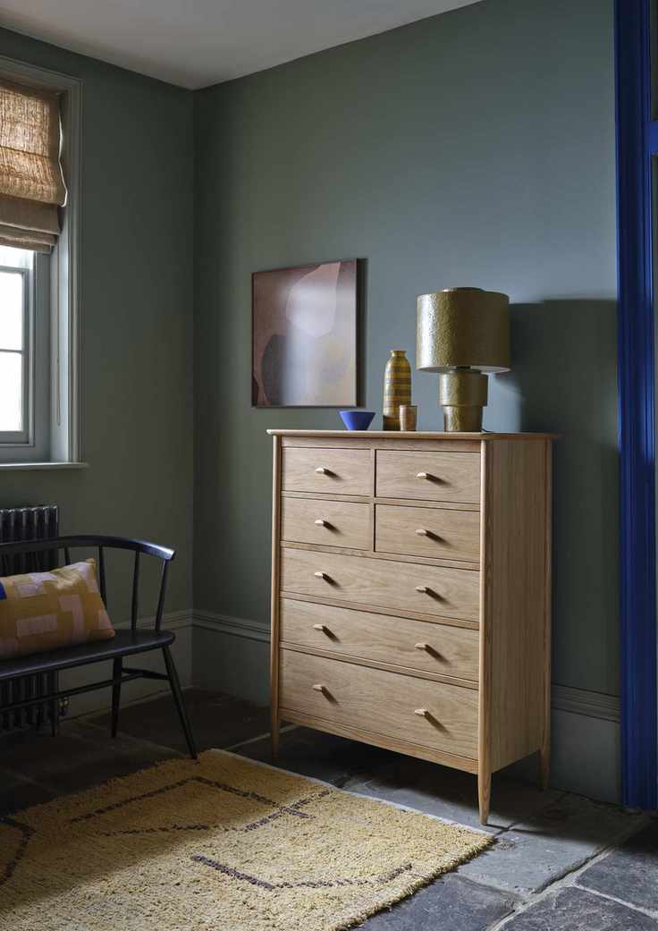 green bedroom with tallboy, stone floor, rug, lamp, bases, artwork, ercol bench and cushion, blind at window, electric blue door frame