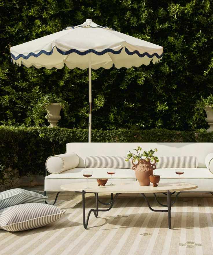 patio area surrounded by green hedges with black metal furniture, white cushions, striped throw pillows, and a white and black scalloped umbrella