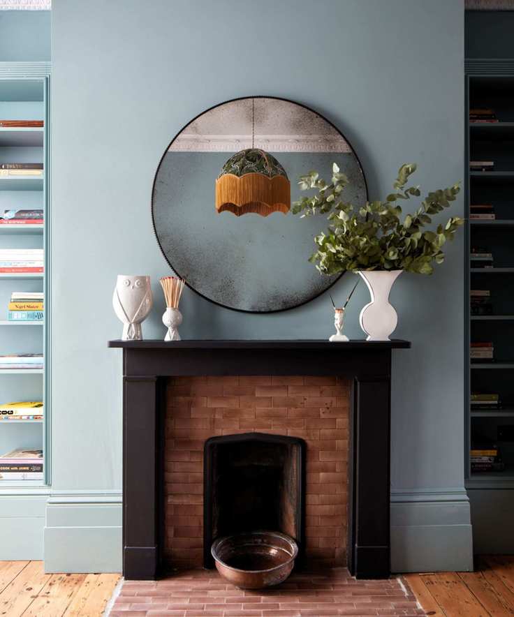 Blue painted wall with fireplace and book shelves and large round mirror. Small vases and ornaments on mantel