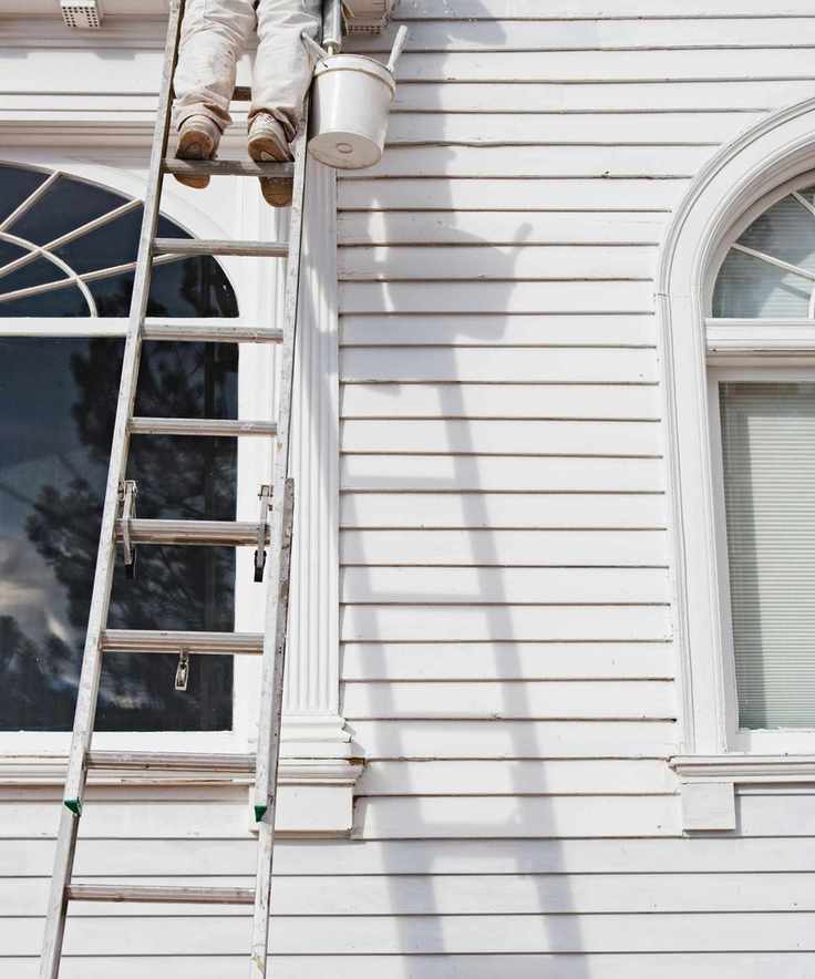 A person standing on a ladder painting the side of a house white