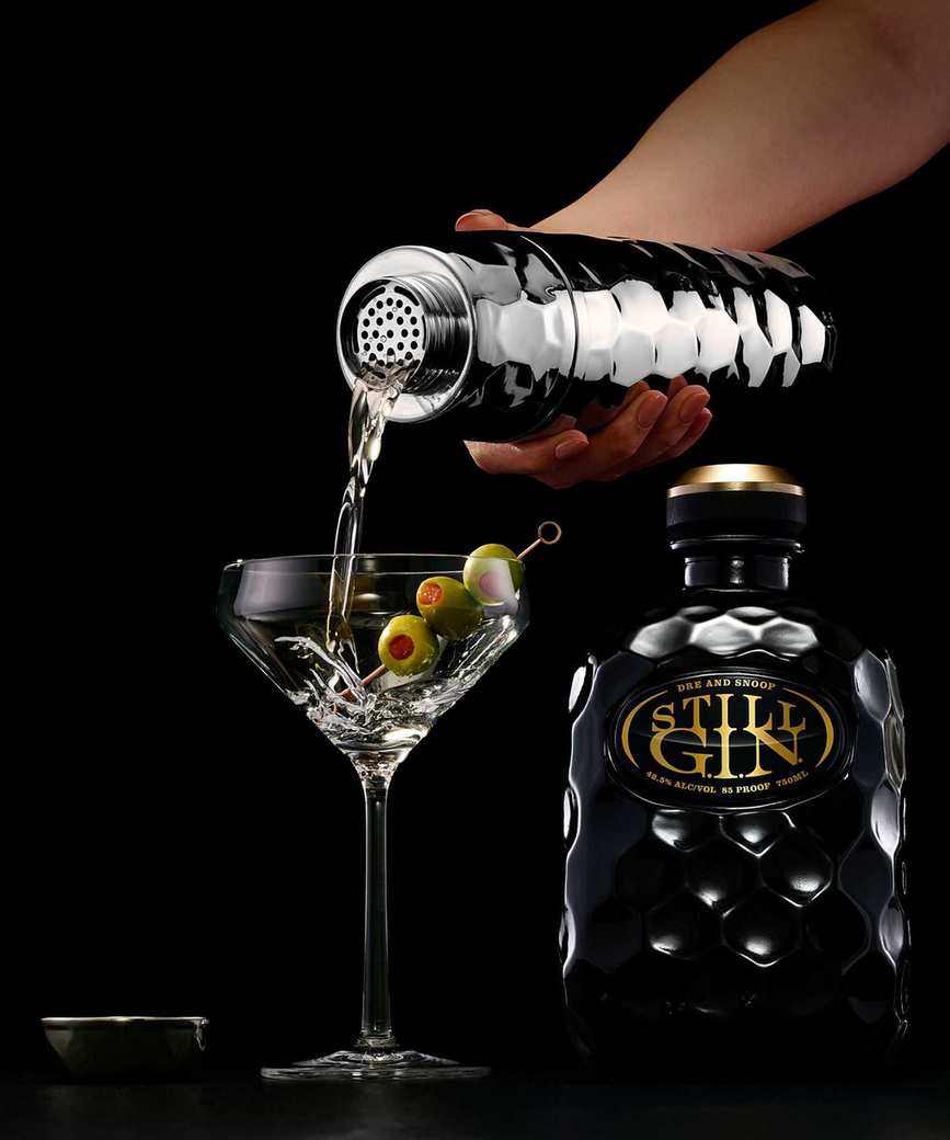Hammered silver cocktail shaker pouring a dirty martini, with a bottle of STILL gin beside it