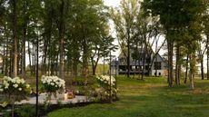 A wide shot of a modern, multi-story farmhouse-style home nestled behind a grove of tall trees, featuring a manicured green lawn and white flowering hydrangeas in the foreground.
