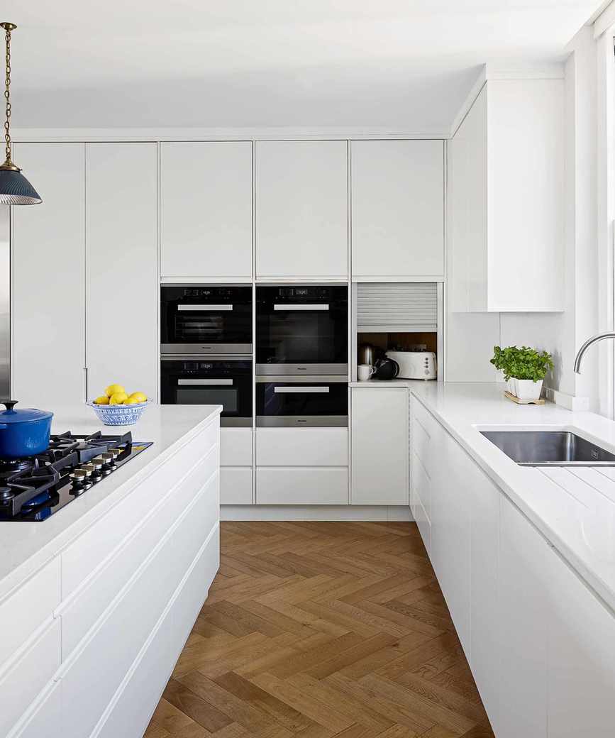 A white kitchen with brown parquet flooring, black double oven and black hobs on a white island with a blue bowl of lemons