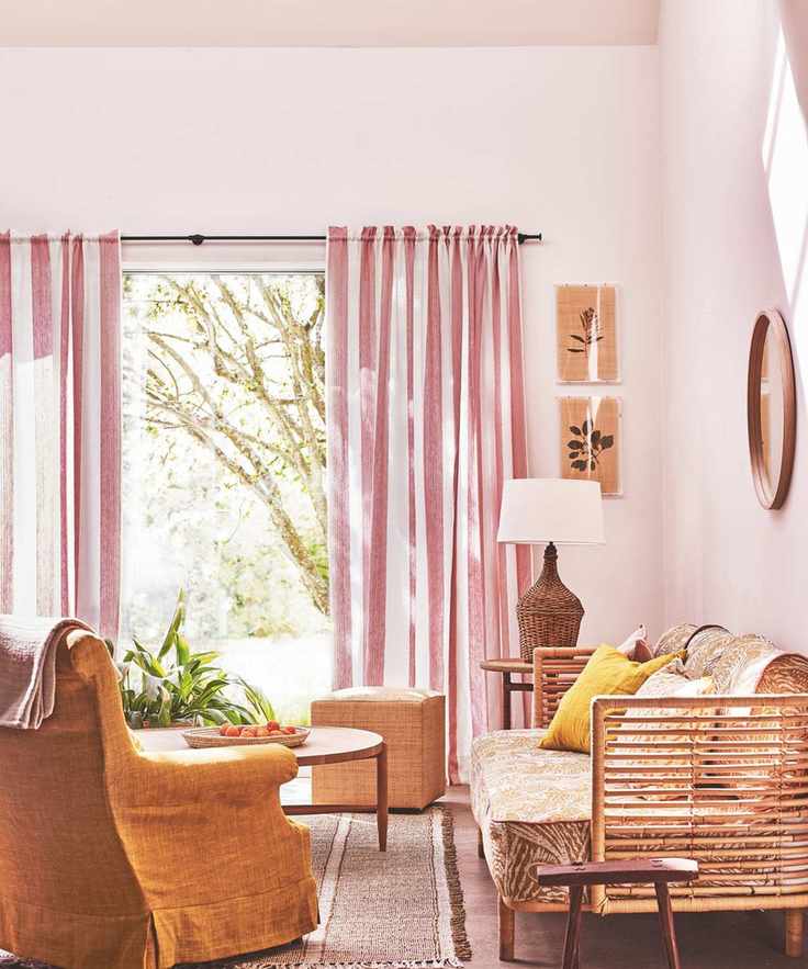 A pink and sunny color drenched living room with a wooden rattan sofa, white and pink striped curtains and a burnt ochre chair