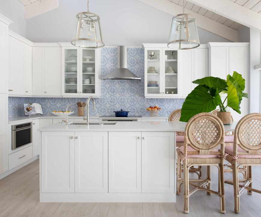 Coastal kitchen with blue and white tiles, white cabinets, white island, rattan bar stools, glass pendants and large palm plant