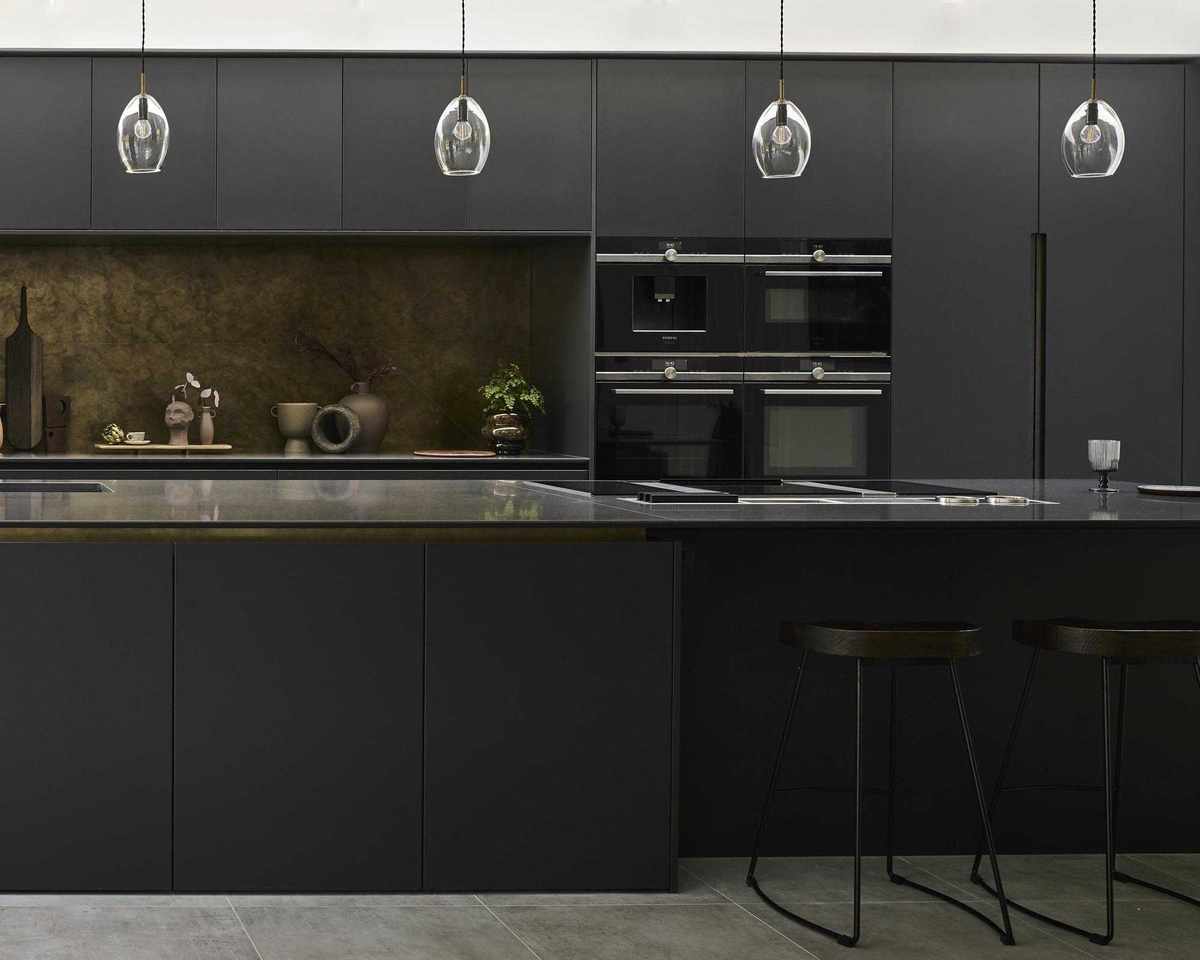 A black kitchen idea with matte black cabinets, stone worktop on the island, brass backsplash and pendant lights