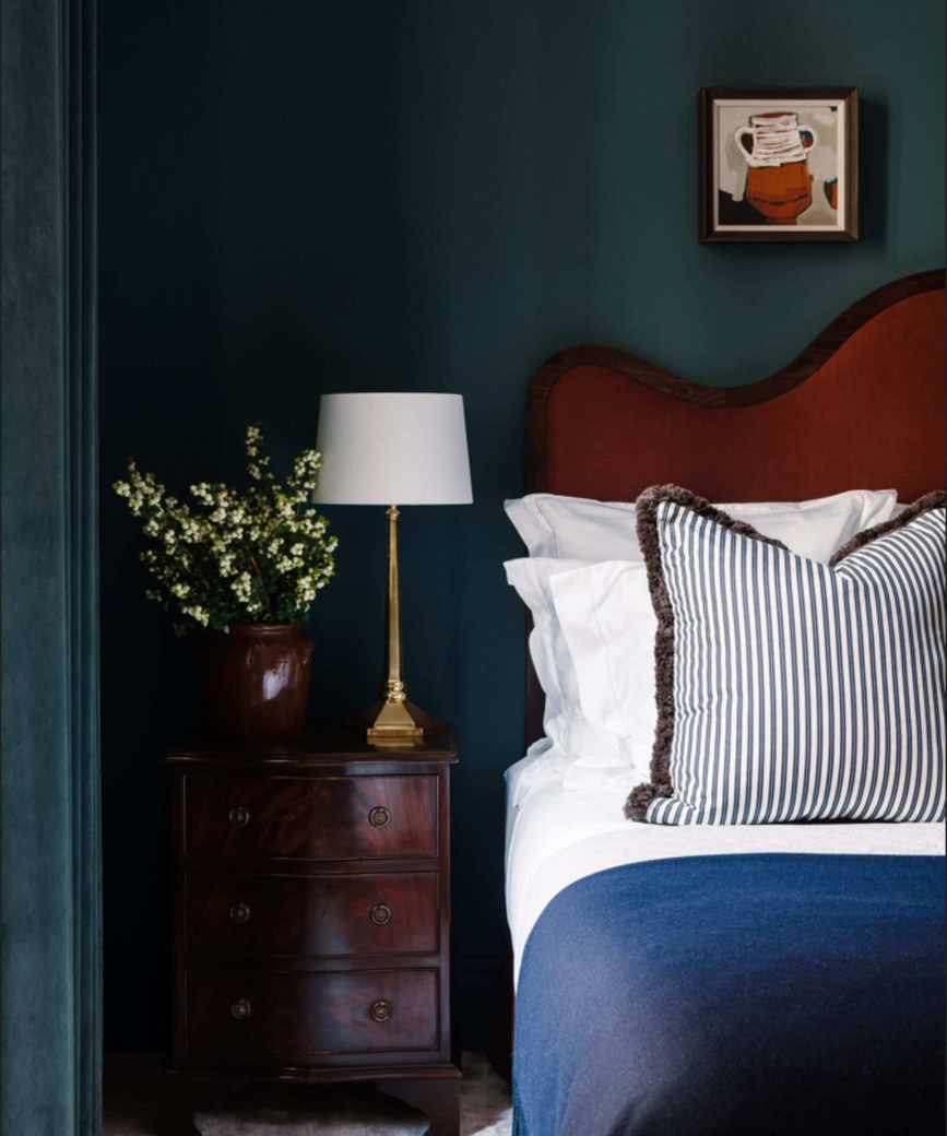 A deep blue bedroom with a red velvet bed, and a dark wood oak side table.