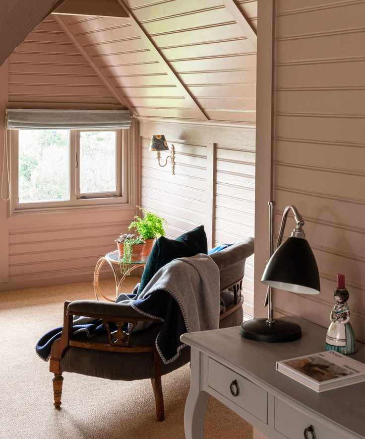 A pink attic room with horizontal painted wood paneling and a blue chair looking out of a window