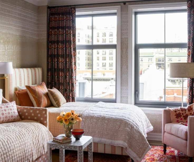 Girls' bedroom with wallpapered walls, printed curtains, striped headboard, floral orange and pink bedding, side table and armchair