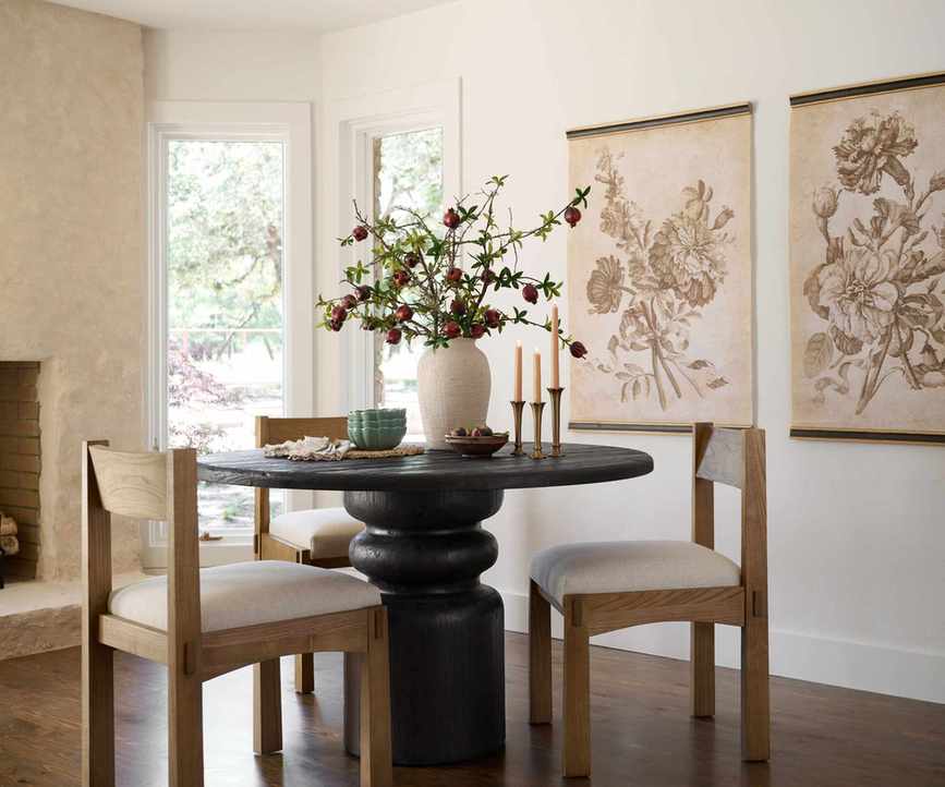 Dining room with curved dark wood table, light wood dining chairs, vintage prints on walls of flowers, large white vase with flowers and gold candlesticks