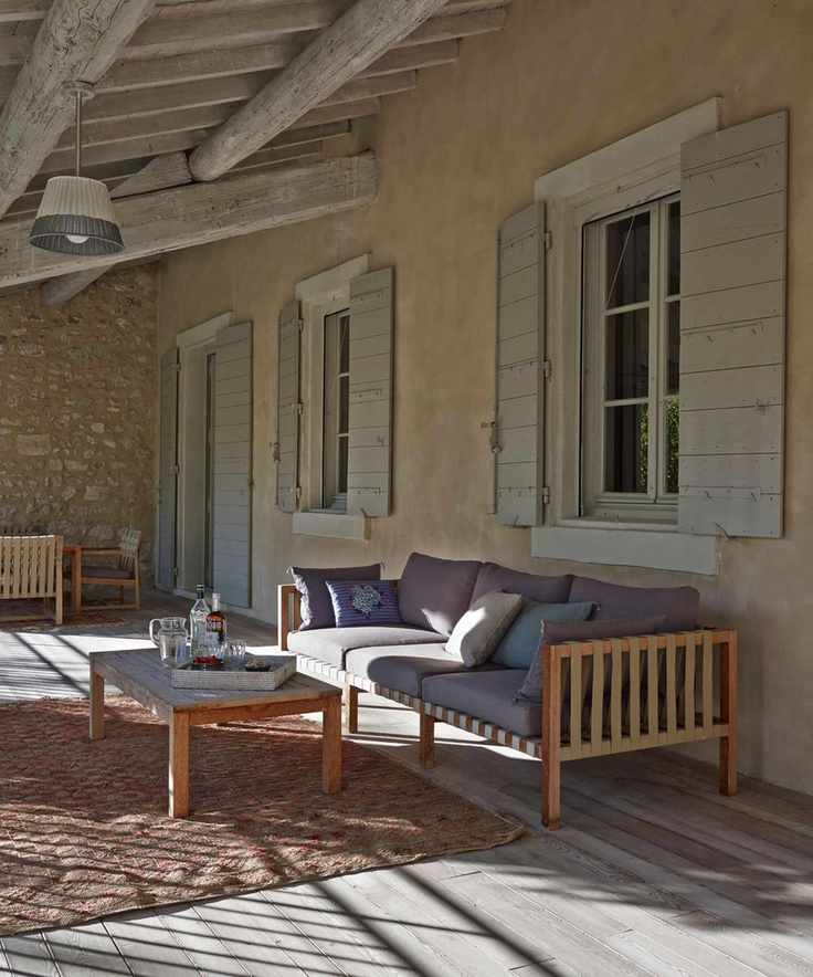 A large porch seating area with shuttered windows, a large wooden sofa with purple cushions, wooden coffee table and large outdoor rug. Above are wooden beams and a light fixture with a white and grey lampshade.