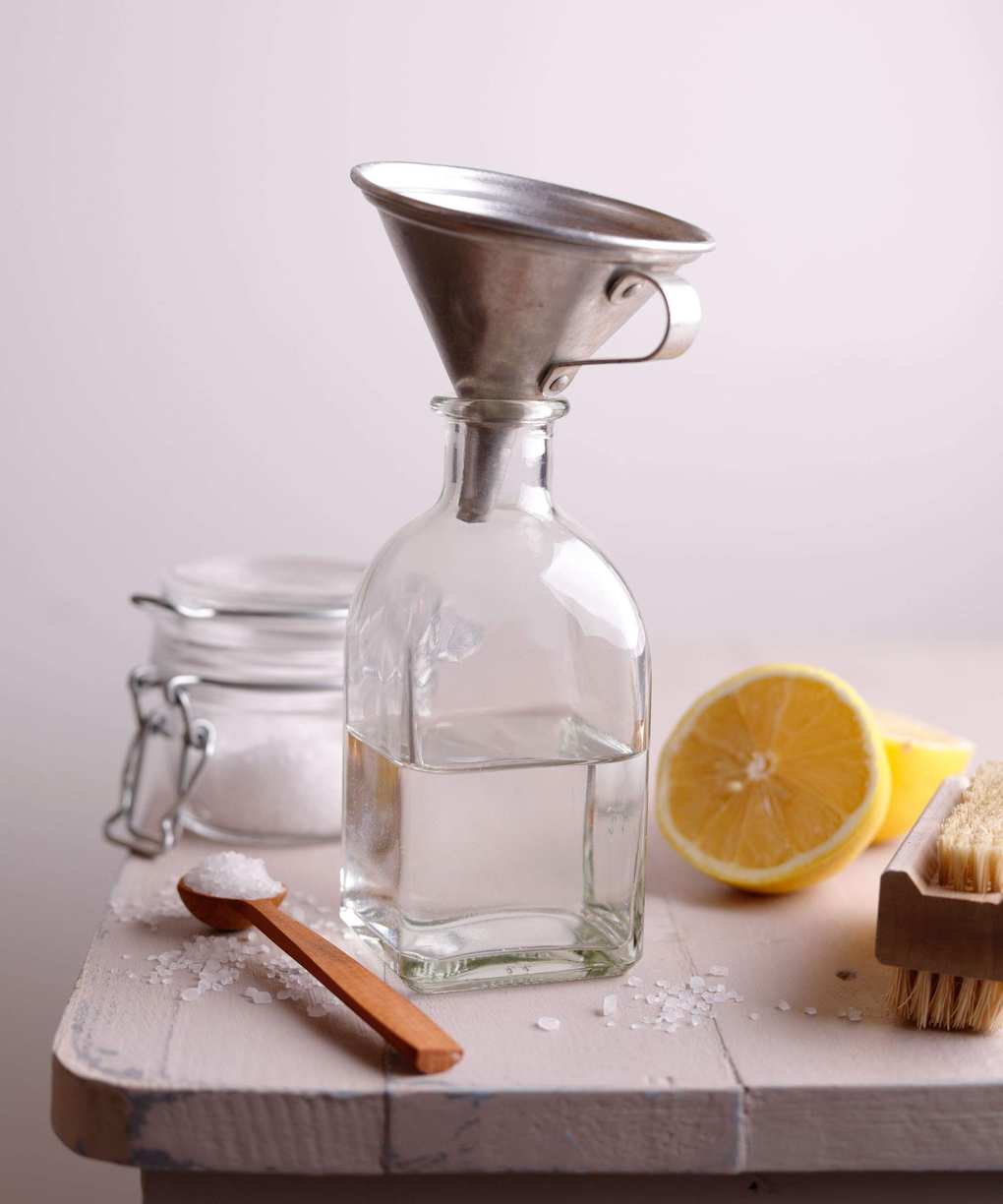 A small glass bottle filled with white vinegar, a metal funnel in the top. A wooden teaspoon of baking soda beside it and a half-cut lemon. On a wooden table.