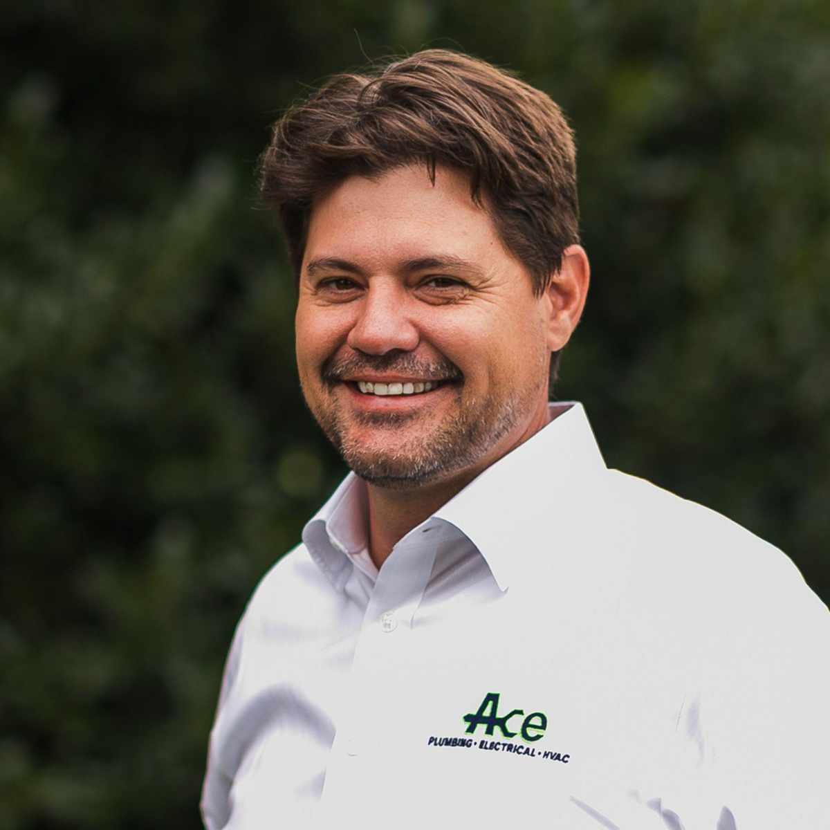 A headshot of Justin Cornforth - a man with brown hair wearing a white shirt, standing in front of dark green shrubbery