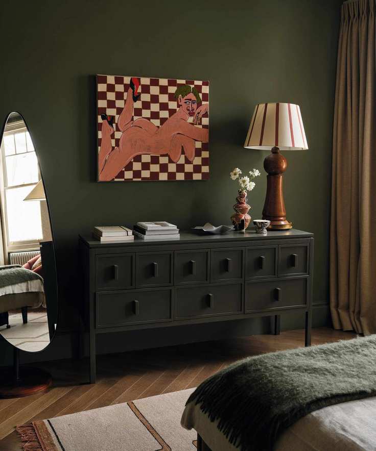 a dark green primary bedroom with abstract artwork on the wall, a large credenza, freestanding mirror and a wooden and striped lamp