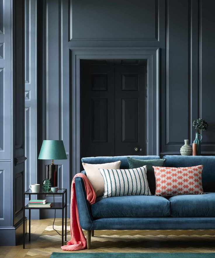 Blue painted living room with paneling, blue sofa with colorful scatter cushions and pink throw, metallic side table with blue table lamp, light wood flooring