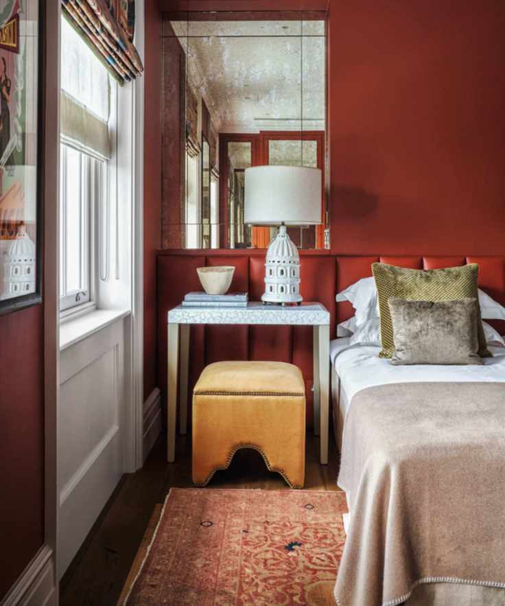 A wall to wall upholstered headboard in a bedroom painted red with a small mustard yellow stool