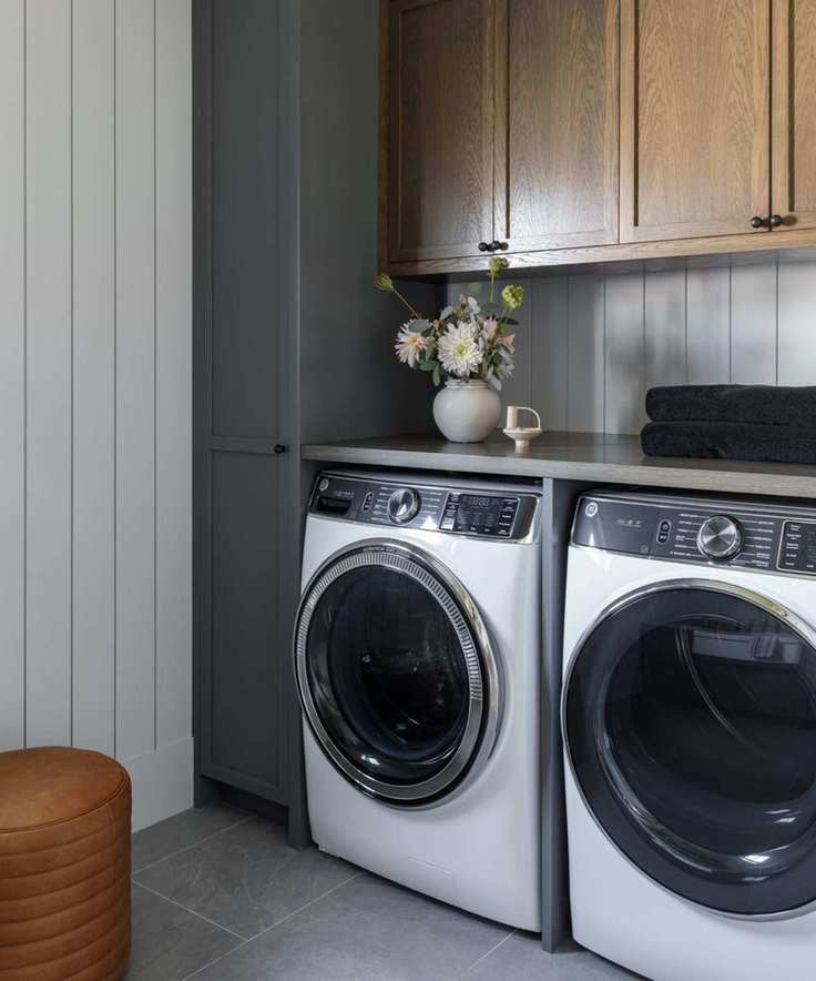 A utilitarian and very clean, uncluttered laundry room. Visible is a counter with a washer and dryer underneath, with cabinets on top and to the side. Plus, stone floor, paneled walls and a round leather stool