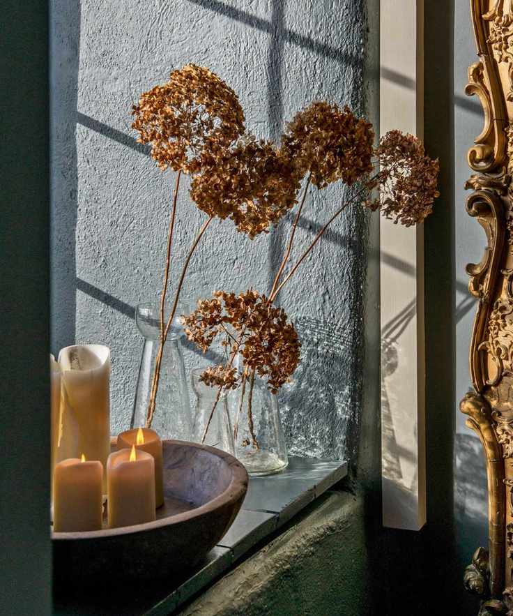 Close up of bowl with candles and dried flowers in glass vase on window sill.