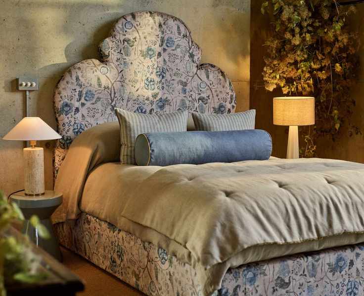 A cozy neutral bedroom with cream and blue floral headboard, mismatched but expensive looking bedside lamps, taupe bedding and blue throw pillows