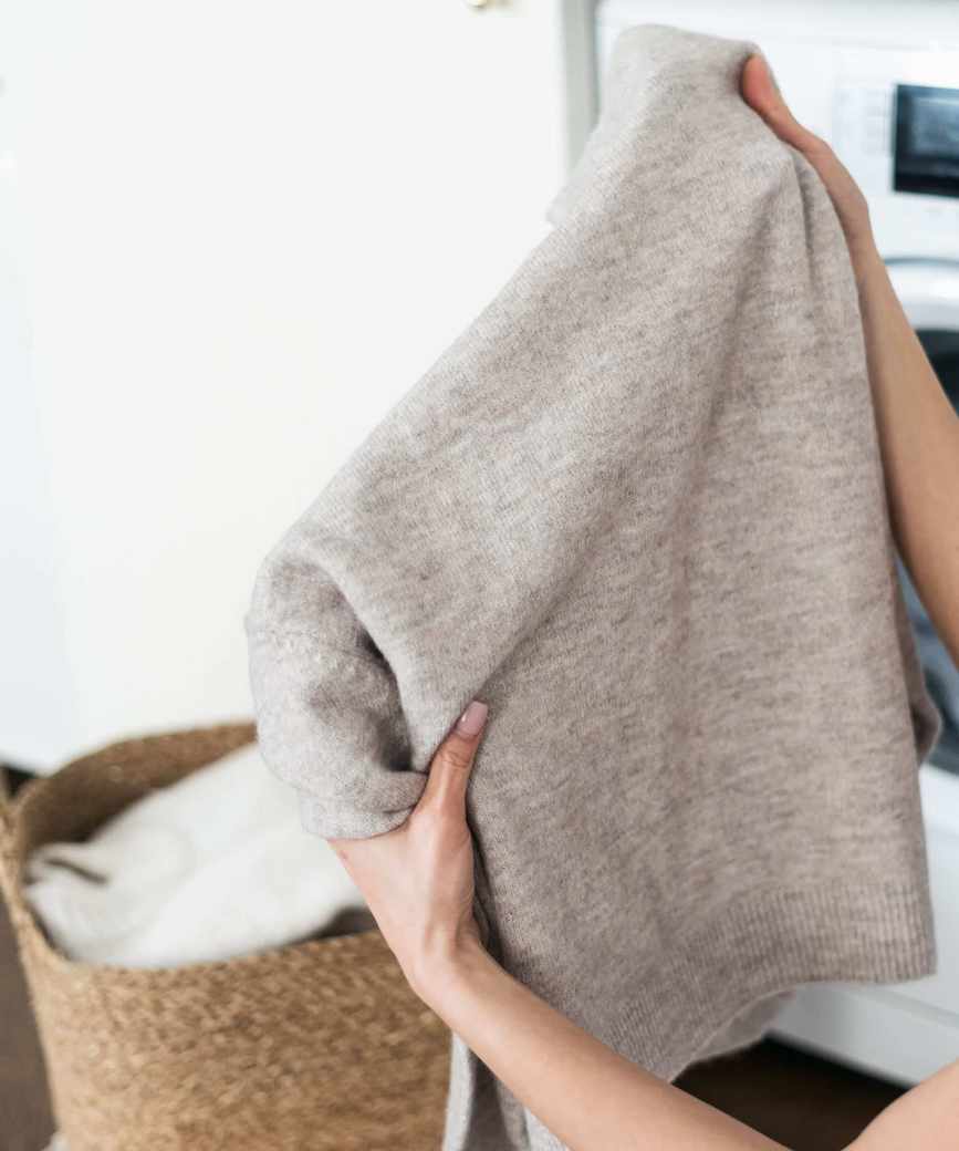 Someone holding up a grey wool sweater in front of a washing machine and laundry basket